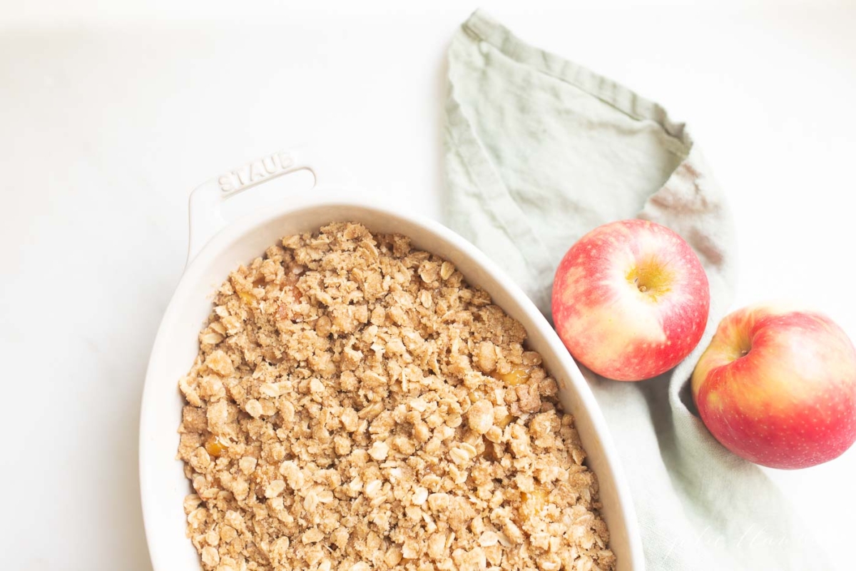 Apple crisp in a white dish surrounded by freshly sliced apples, showcasing its deliciousness.