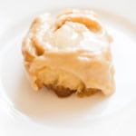 A close-up of a single glazed cinnamon roll with caramel icing drizzled on top, presented on a white plate.