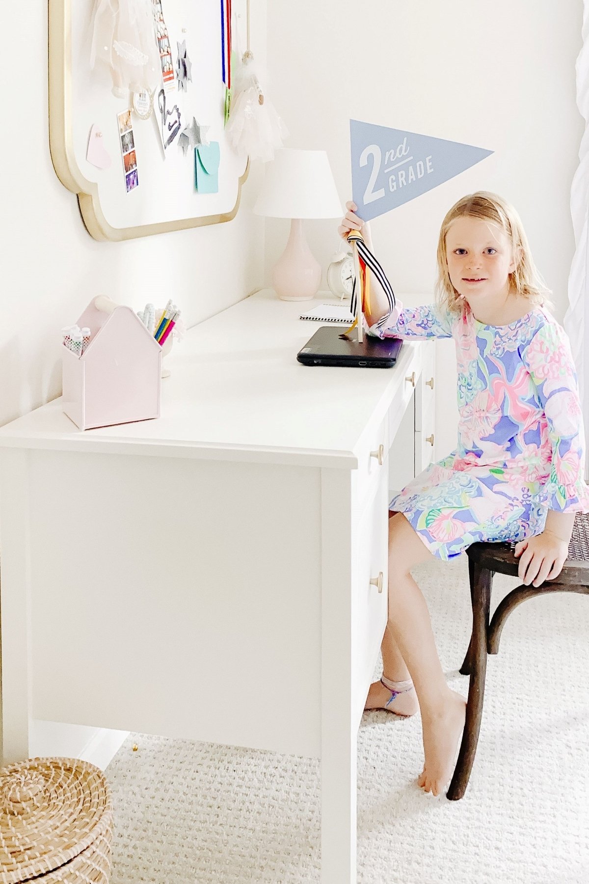 A little blonde girl at a white desk in a white bedroom, holding a flag that says 2nd grade.