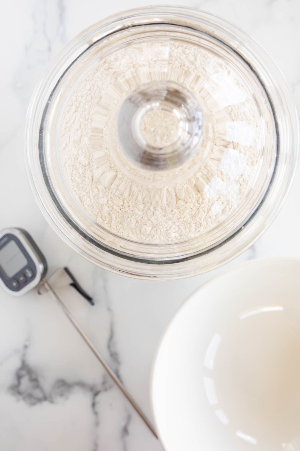 A glass canister full of flour, with a white bowl and a digital thermometer to the side