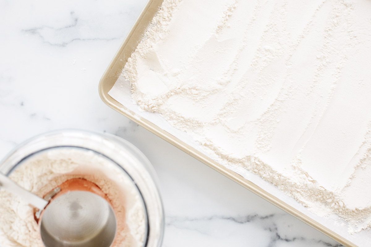 A canister of flour next to flour spread out on a baking sheet, all placed on a marble countertop.