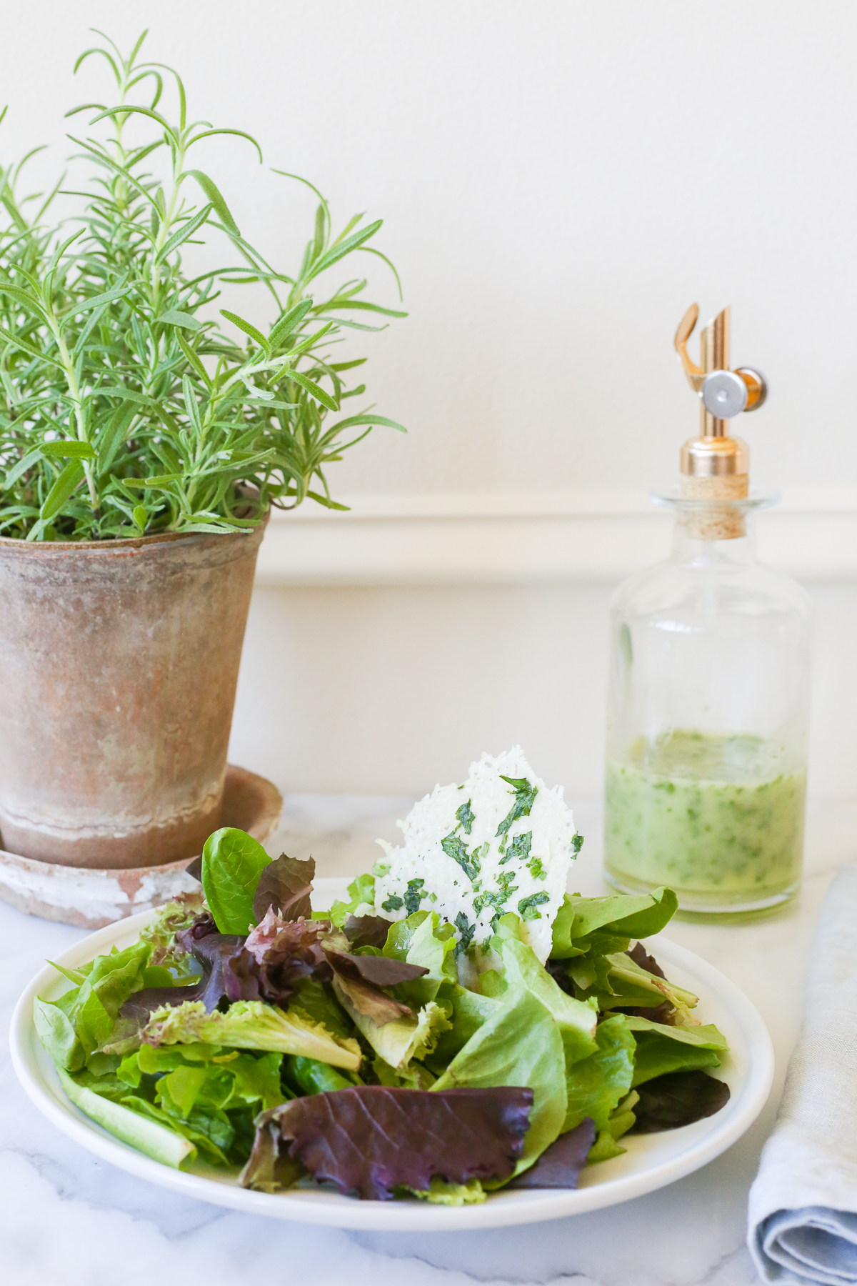 A bottle of homemade vinaigrette next to a plate of salad and a pot of rosemary 