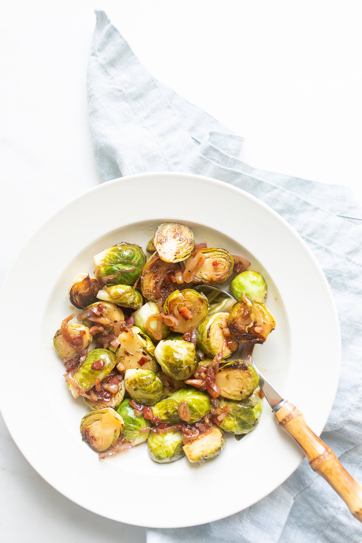 A white plate with cooked Brussels sprouts, a perfect meatloaf side, garnished with caramelized onions and garlic on a light blue cloth. A fork with a wooden handle rests on the plate.
