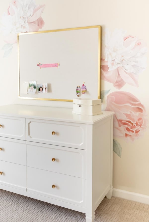 A girl's bedroom with cream walls and peony wallpaper behind a white dresser.
