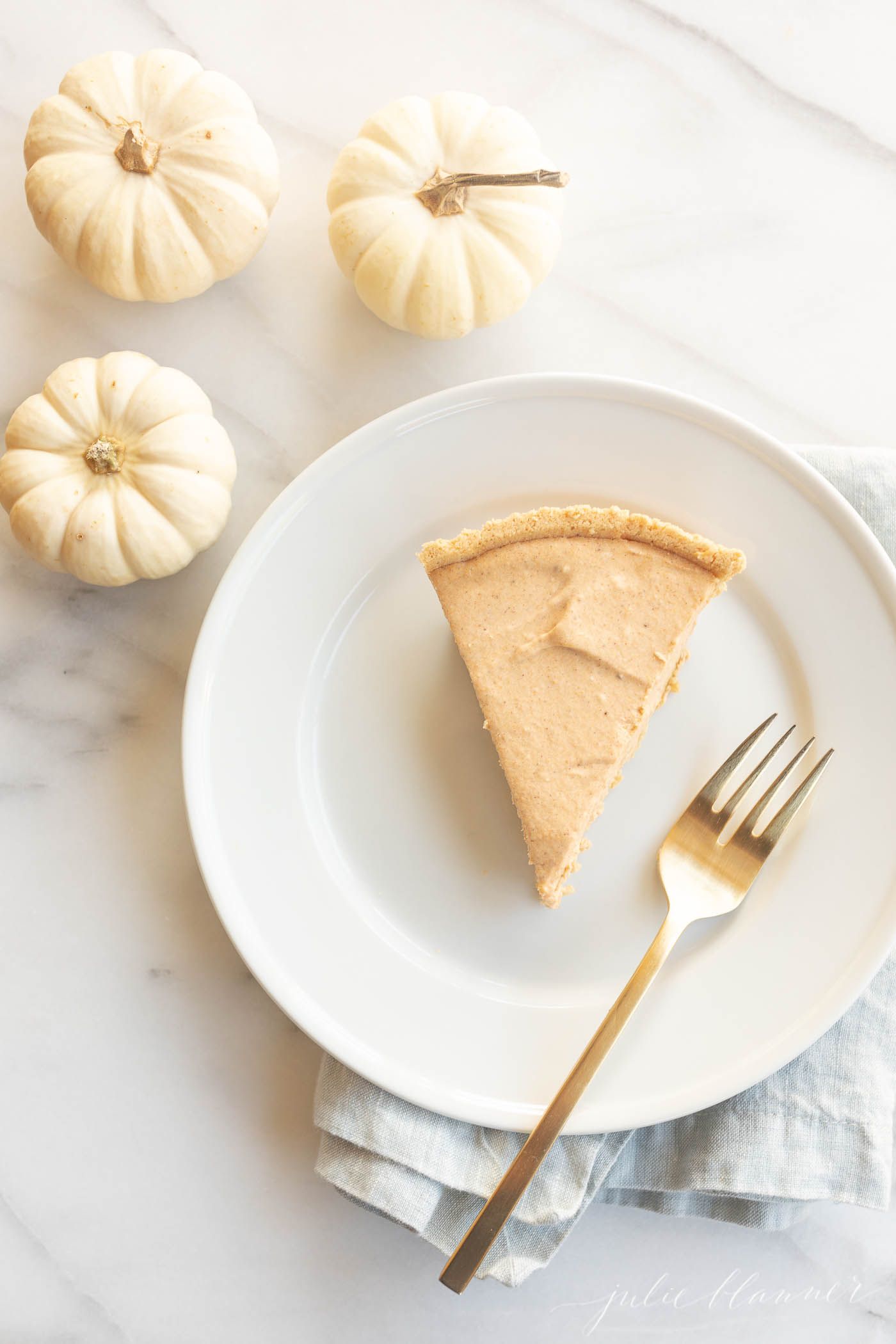 A slice of pumpkin cheesecake on a white plate. Three tiny white pumpkins are in the background, and the whole scene is placed on a marble countertop.