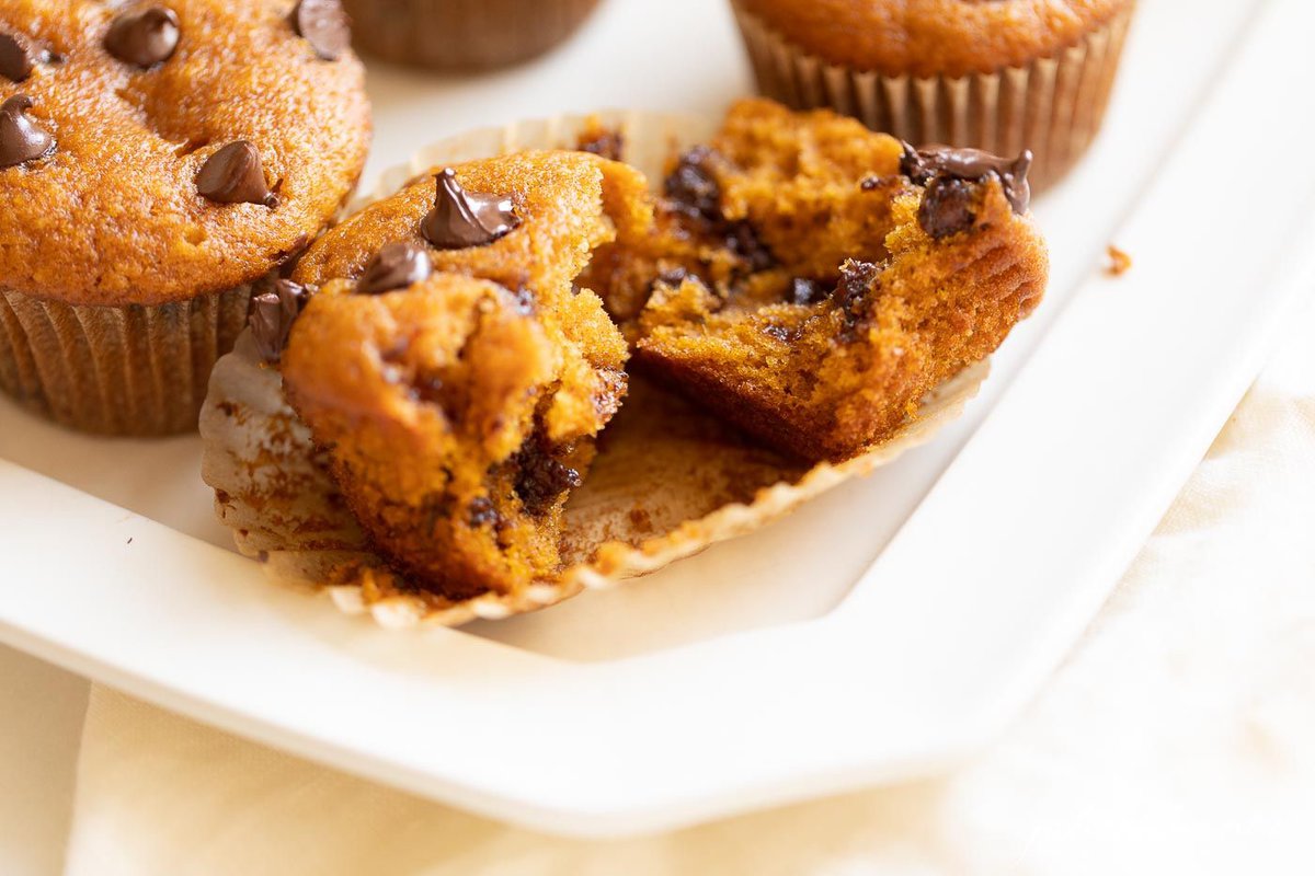Pumpkin chocolate chip muffins on a white platter, the first one split into two pieces in a wrapper.