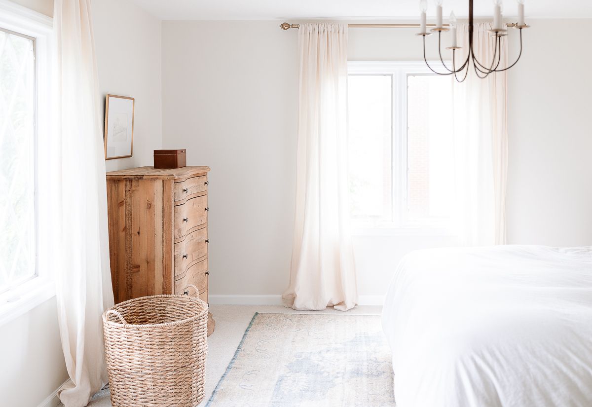 A soft blue antique rug on the carpet in a white bedroom, a wooden chest of drawers in the left corner.