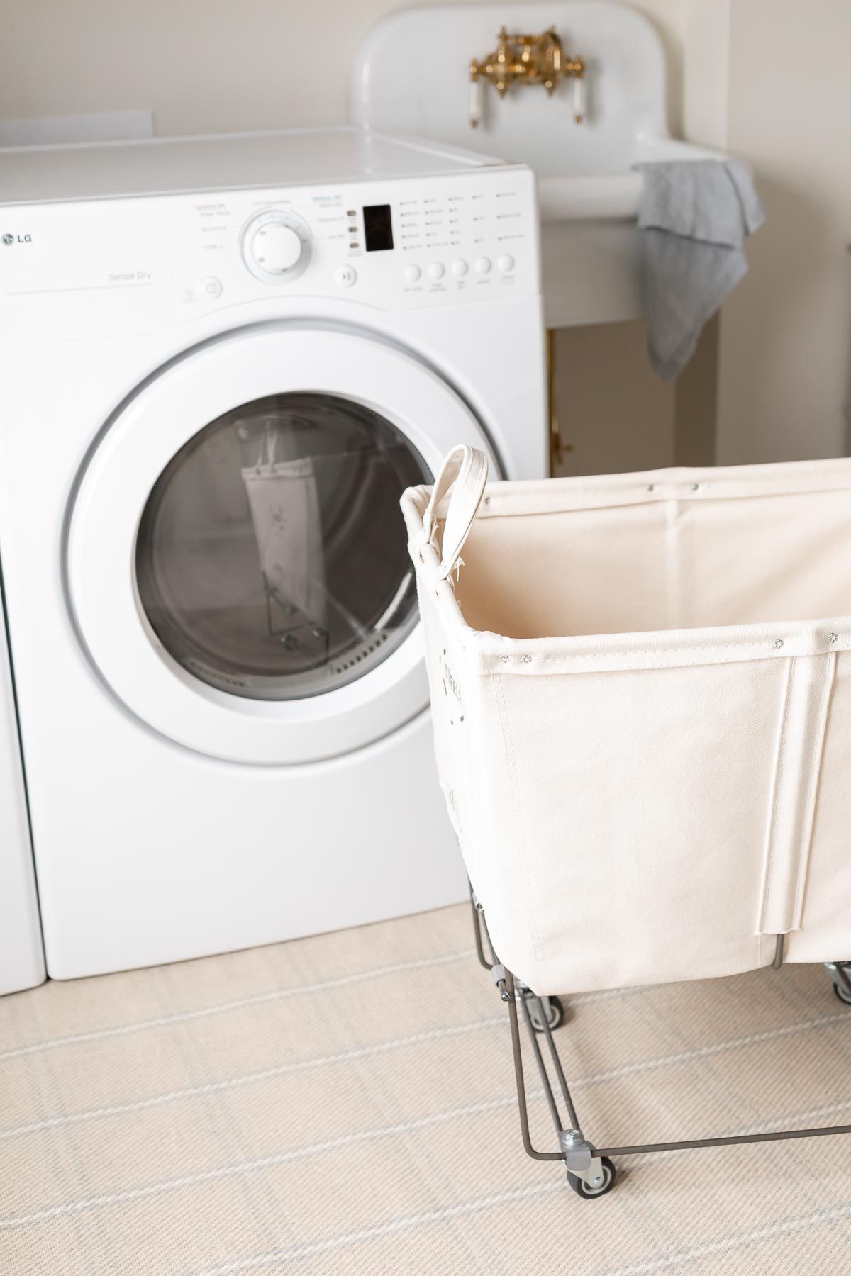A laundry room with a white tumble dryer, a vintage laundry basket in front, placed on top of plaid rugs on the carpet