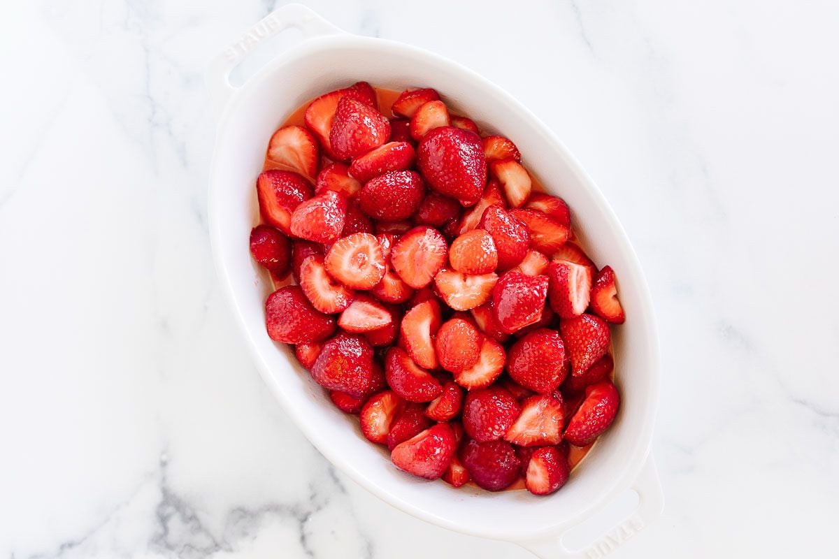 An oval white baking dish filled with fresh sliced strawberries, placed on a marble surface