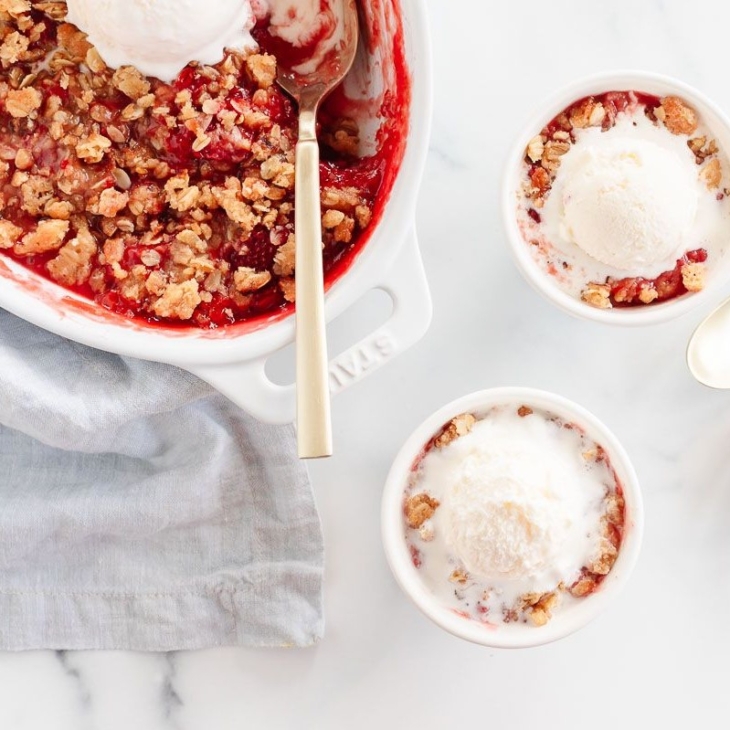 A strawberry crumble dessert in an oval baking dish, two servings in white bowls topped with ice cream to the side.