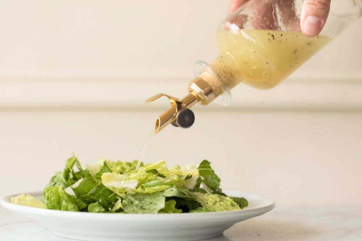 A hand pouring white wine vinaigrette from a glass bottle onto a plate of green leafy salad placed on a white surface.