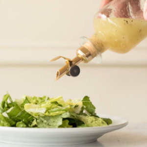 A person pours white wine vinaigrette from a glass bottle with a brass spout onto a plate of fresh green salad.