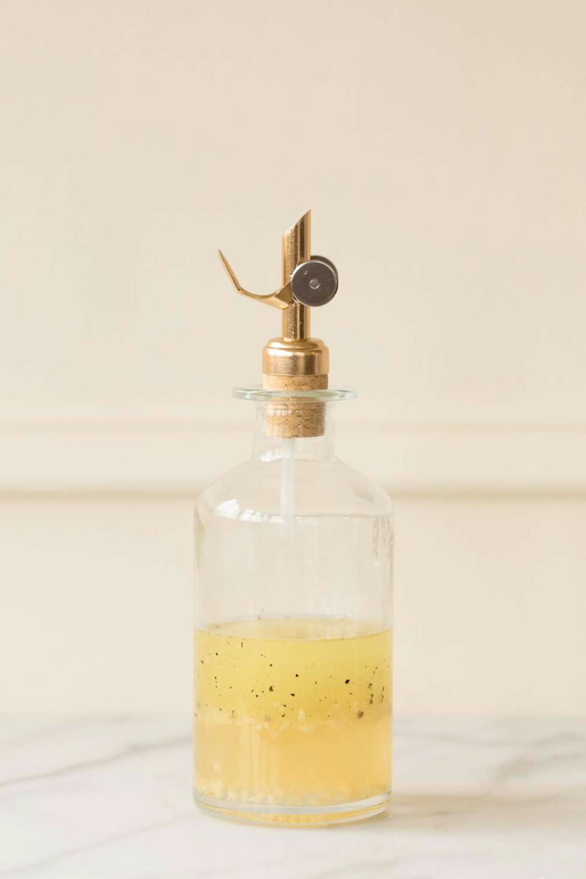 Clear glass oil dispenser with a metal spout, partly filled with a yellow liquid containing black specks—resembling a homemade white wine vinaigrette—placed on a white surface against a light background.