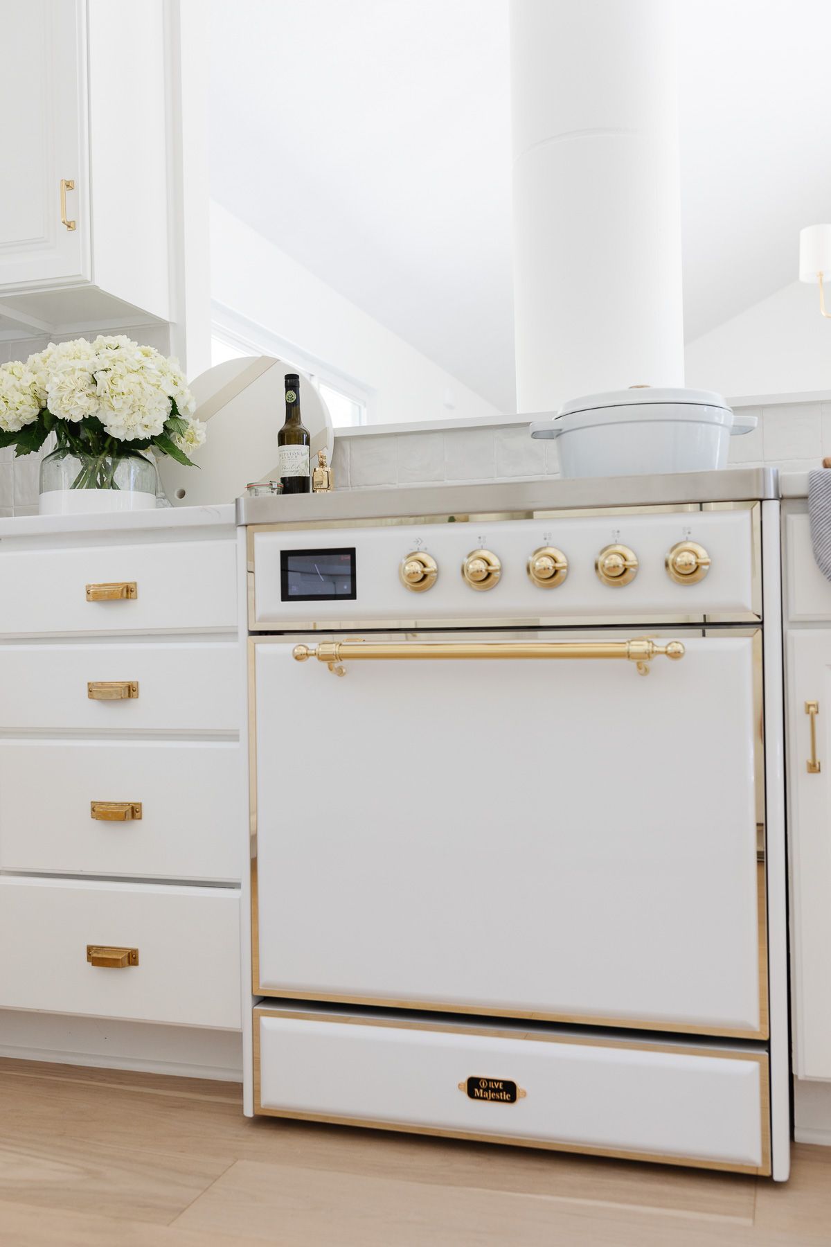 A close up of a white ILVE range in a kitchen with white cabinets and brass hardware