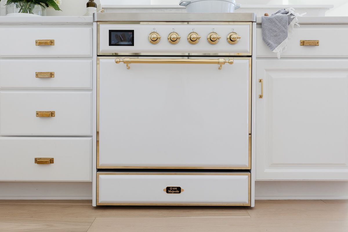 A close up of a white ILVE range in a kitchen with white cabinets and brass hardware