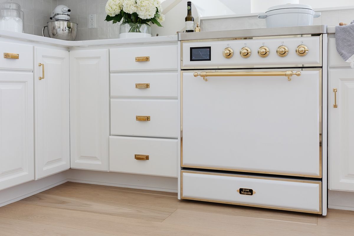 A close up of a white ILVE range in a kitchen with white cabinets and brass hardware