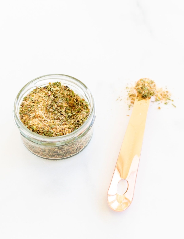 A small glass container filled with all purpose seasoning, teaspoon to the side.
