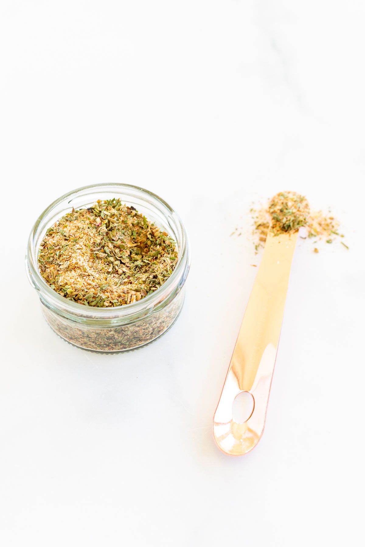 A small glass container filled with all purpose seasoning, teaspoon to the side.