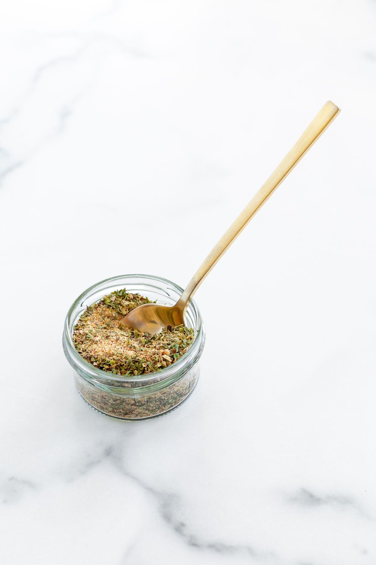 A small glass container filled with all purpose seasoning, gold spoon in the jar.