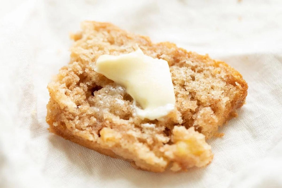 A close-up of a partially eaten cinnamon crumb muffin with a small pat of melted butter on top, reminiscent of an apple bread recipe, resting on a white cloth.