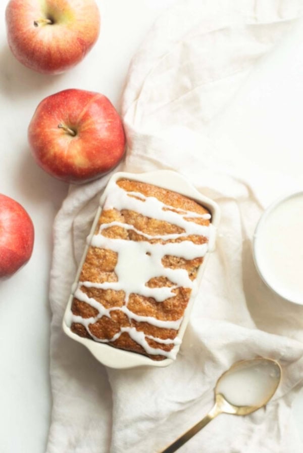 A loaf of apple bread with white icing drizzled on top, surrounded by three apples. A spoon and a bowl of icing are placed on a white cloth beside the bread, inviting you to try this delightful apple bread recipe.