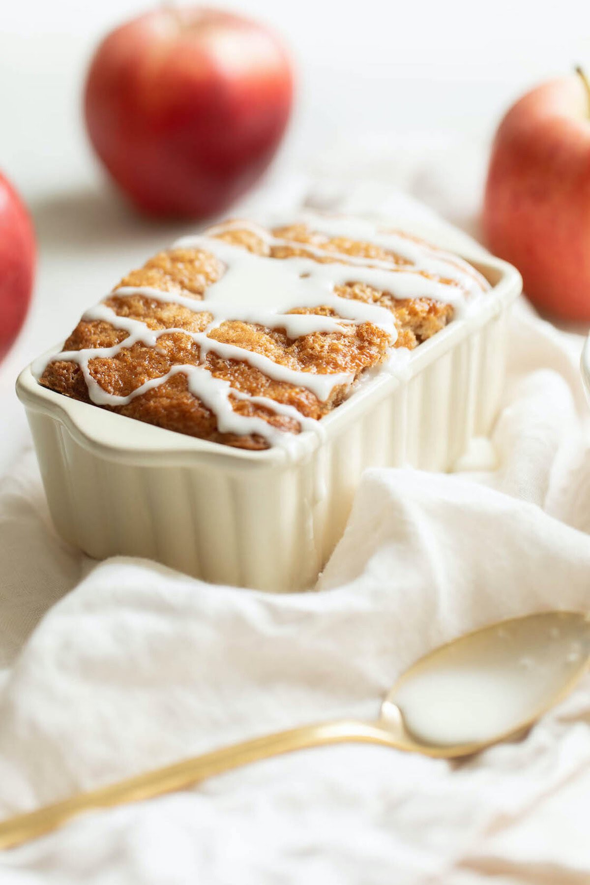 A glazed apple bread recipe in a rectangular baking dish is set against a white cloth, with two apples and a spoon nearby.