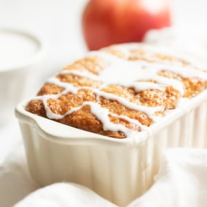 A freshly baked loaf cake, reminiscent of a classic apple bread recipe, is topped with white icing in a rectangular ceramic dish. An apple and a small bowl subtly blur into the background, enhancing the cozy kitchen vibe.