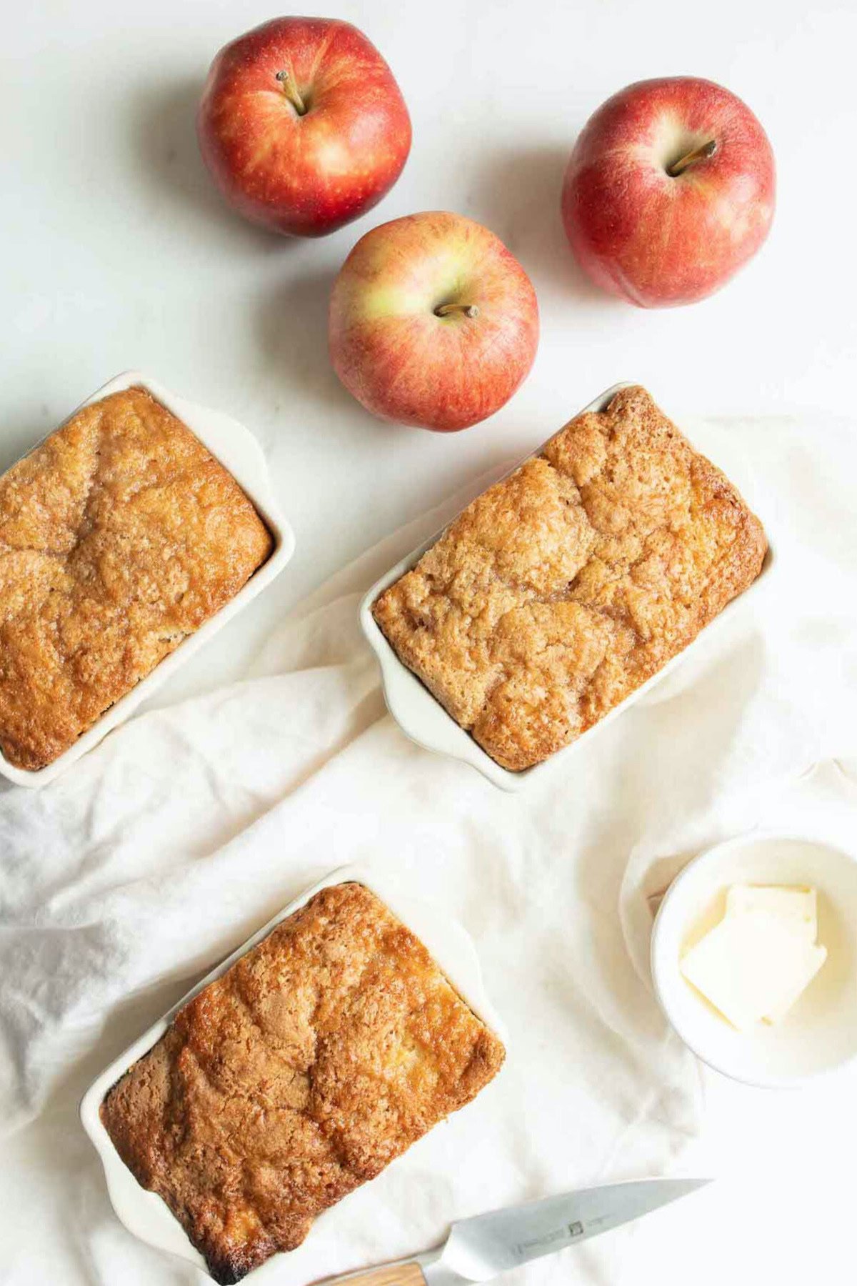 Three baked apple bread loaves in white dishes rest on a white cloth, accompanied by three red apples and a butter dish. A knife with a wooden handle peeks into the frame at the lower right corner, showcasing the perfect scene for an apple bread recipe.