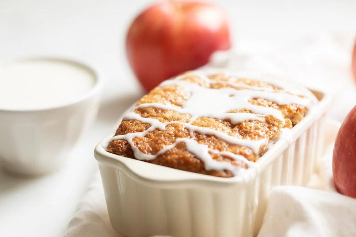 Loaf of glazed apple bread in a white ceramic dish with an apple and a cup of milk in the background, showcasing a delightful apple bread recipe.