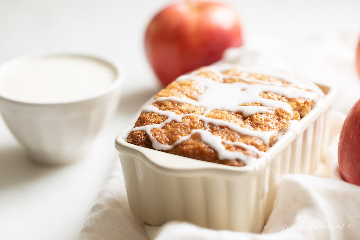 A small loaf pan of apple bread, topped with white icing and surrounded with apples and a linen napkin. 