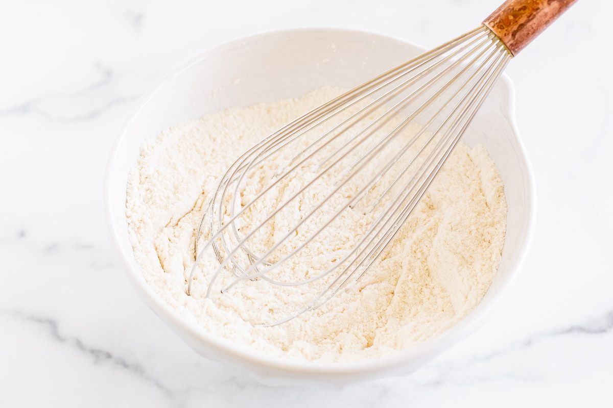 A white mixing bowl with flour and a whisk, placed on a marble countertop