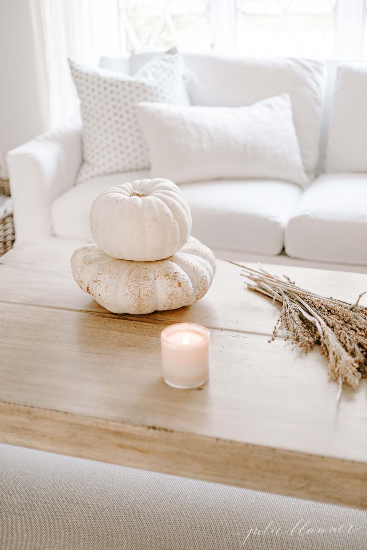 Coffee table featuring a candle and white pumpkins, open basket filled with blankets in background.