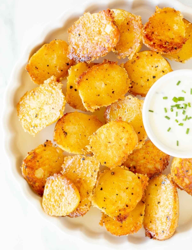 A white ruffled plate full of parmesan potatoes, with a small bowl of sour cream dip.