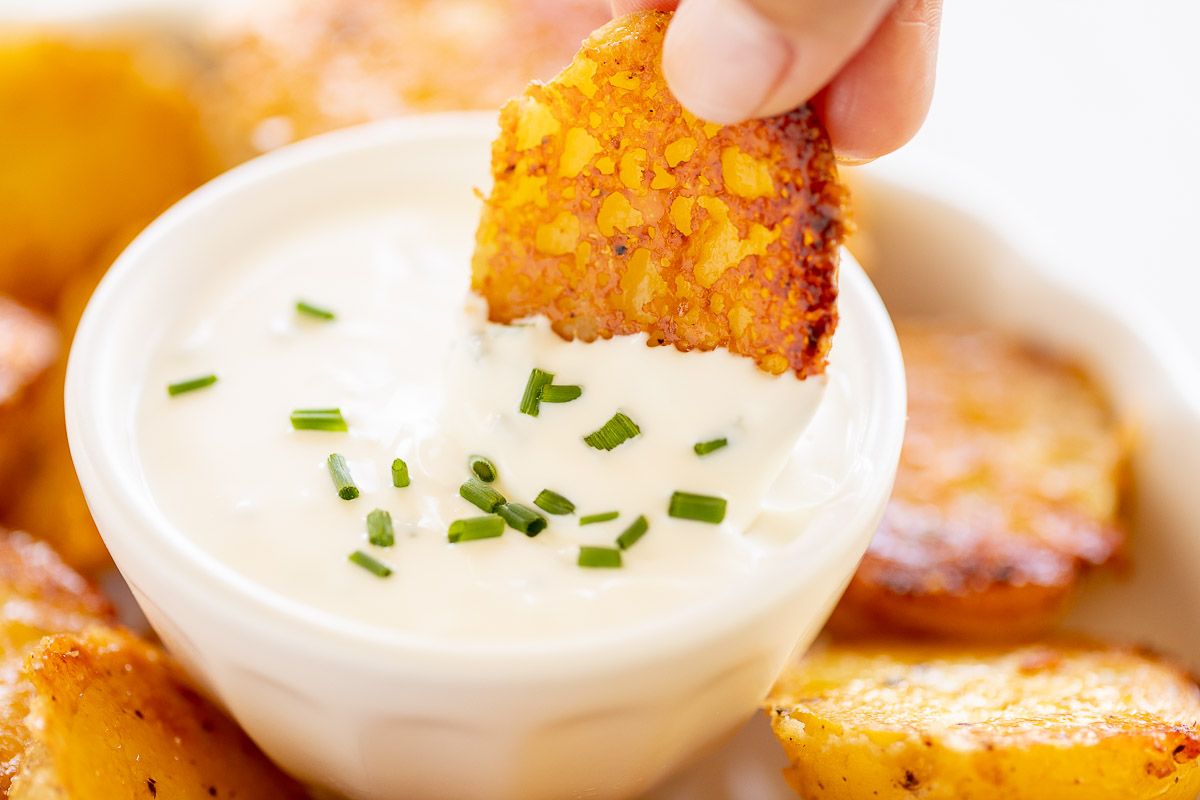 A hand dipping a Parmesan roasted potato into a white bowl of sour cream dip