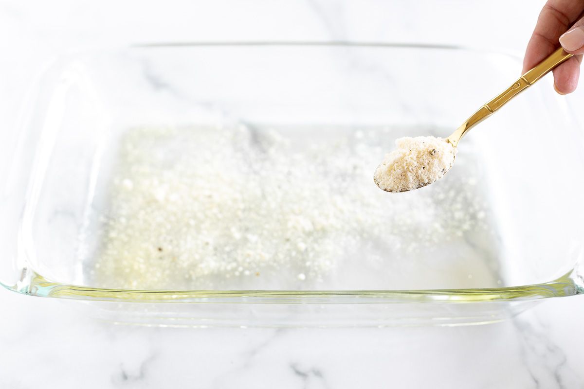 A hand sprinkling a Parmesan mixture onto a clear glass baking dish.