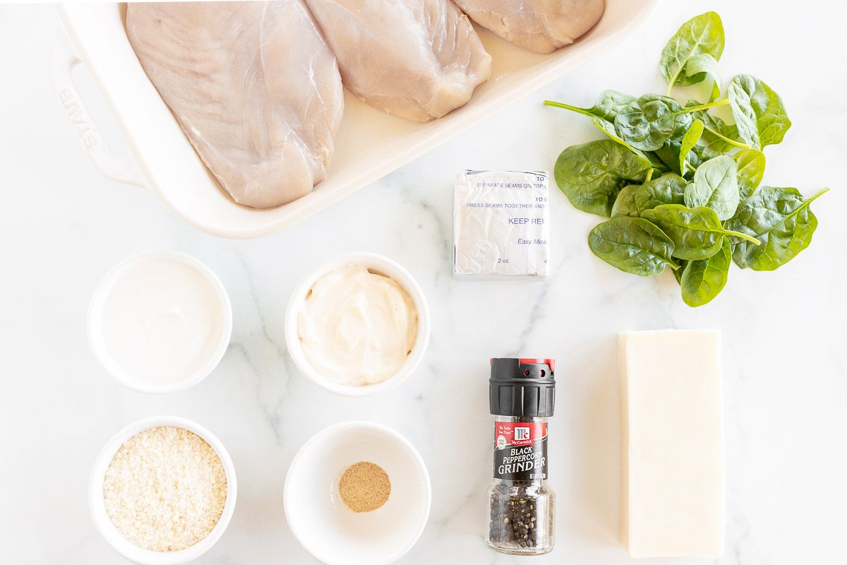 Ingredients for a baked chicken and spinach dish laid out on a white marble surface.