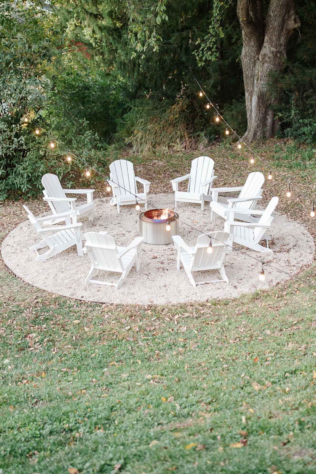 White Adirondack chairs, circling a Solo Stove on a DIY pea gravel fire pit patio. String lights are hung above, twinkling at dusk.