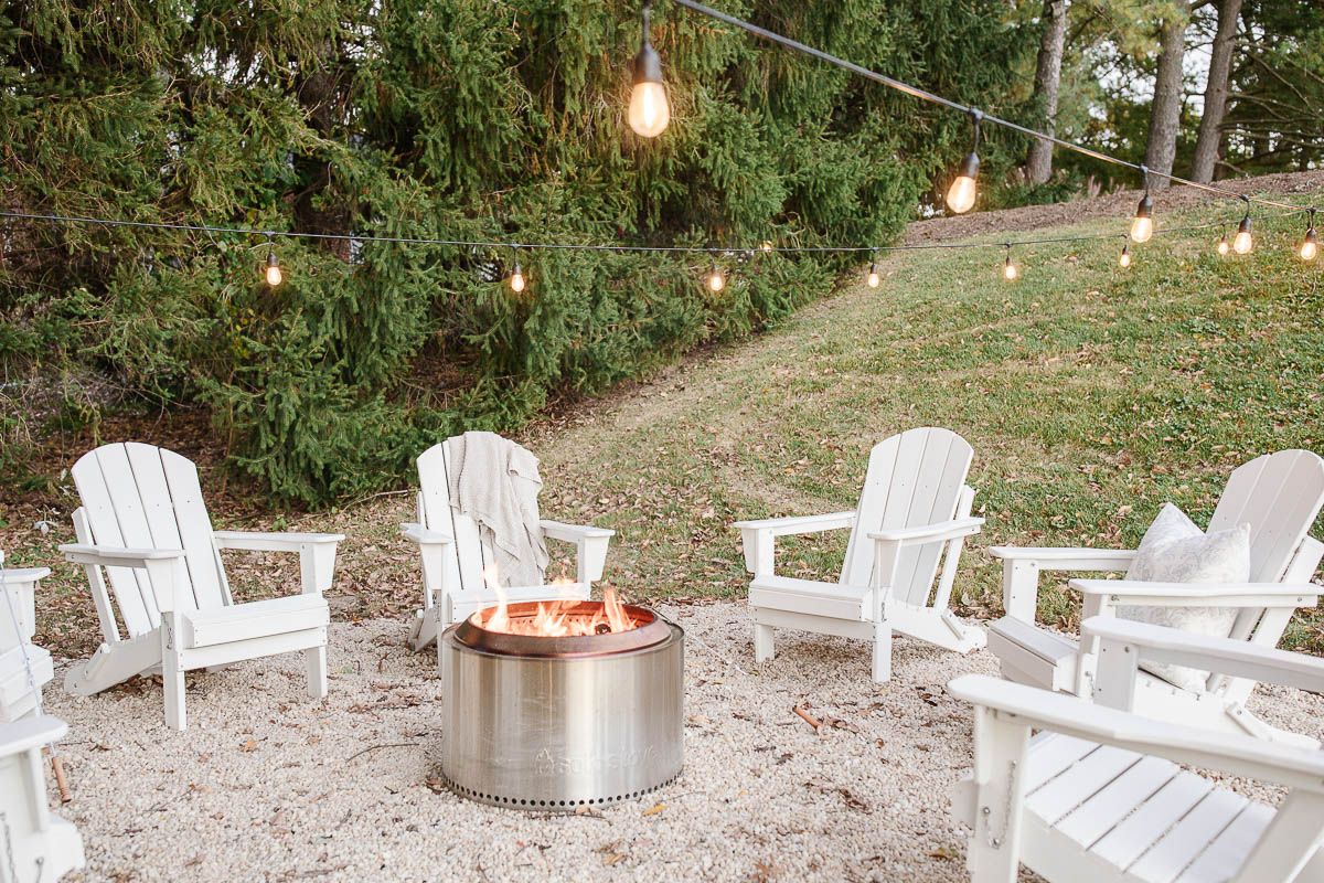 White Adirondack chairs, circling a Solo Stove on a DIY pea gravel fire pit patio. String lights are hung above, twinkling at dusk.