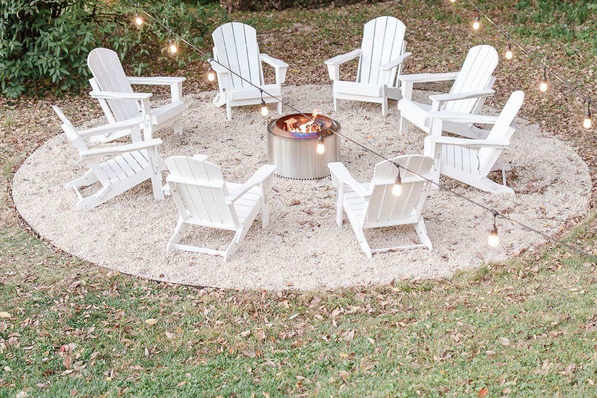 White Adirondack chairs, circling a Solo Stove on a DIY pea gravel fire pit patio. String lights are hung above, twinkling at dusk.