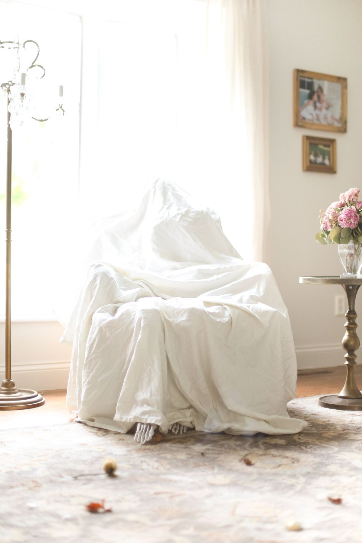 A white living room with Halloween decorating of fall leaves on the floor, and a white sheet over an upholstered chair