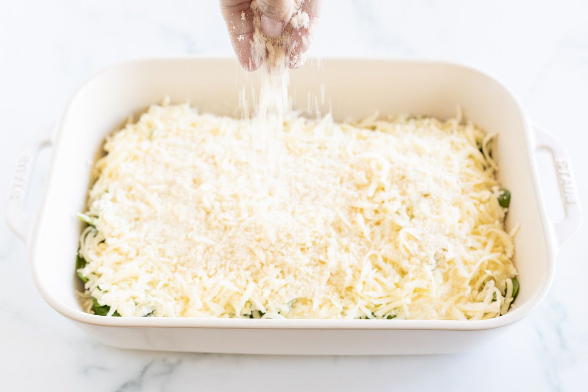 A hand adding cheese on top of a baked chicken and spinach dish.