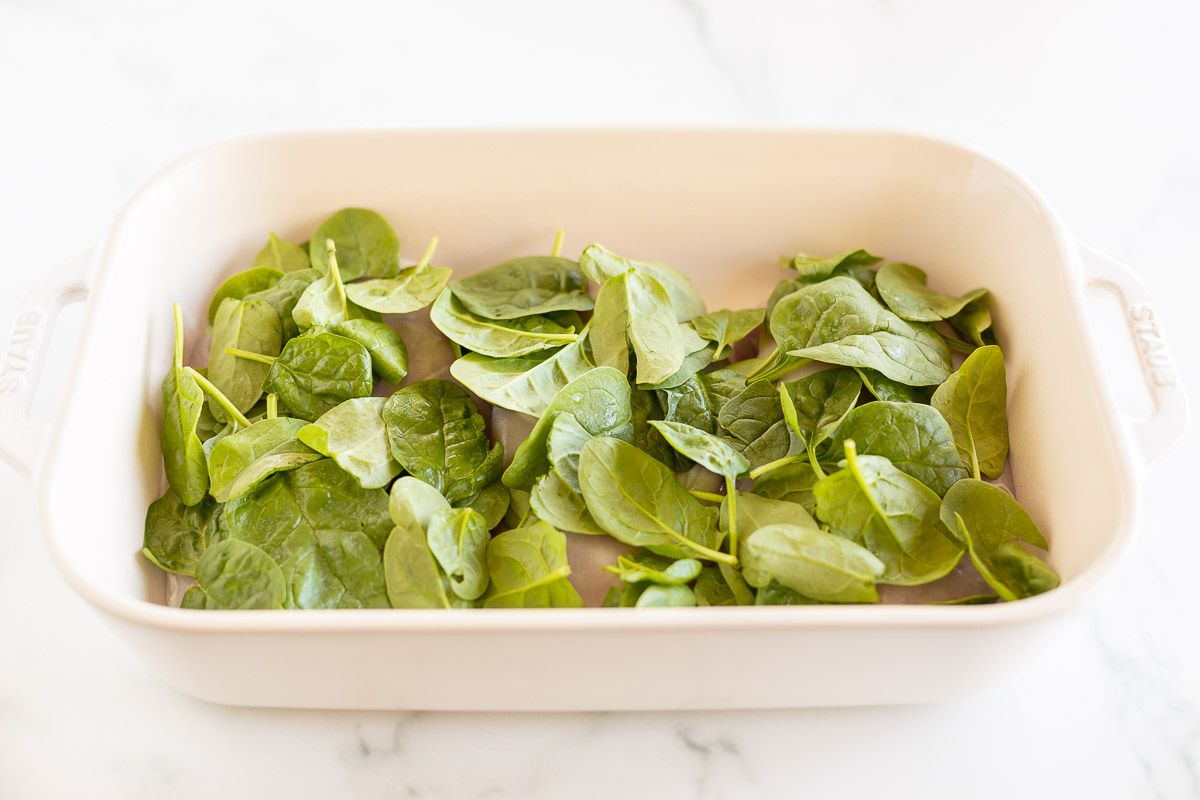 Fresh spinach leaves on top of chicken breasts in a white casserole baking dish.