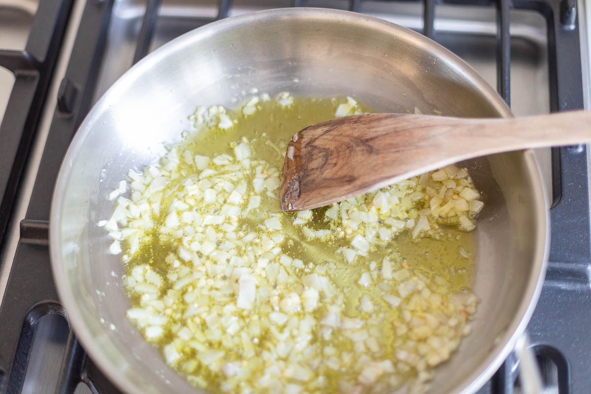 A silver pan on a stove top, wooden spoon stirring shallots and garlic.