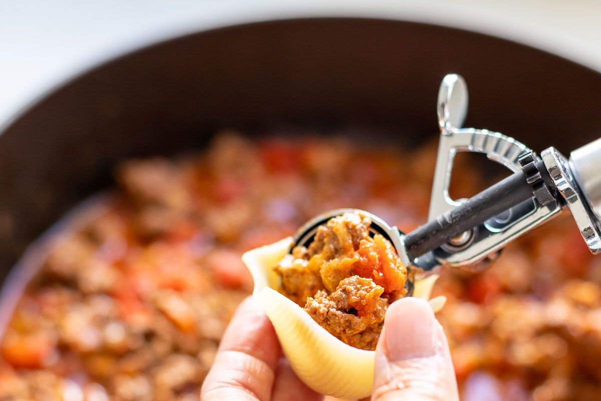 A hand using a cookie scoop to fill jumbo shell pasta with taco meat, pot full of ground beef in the background.