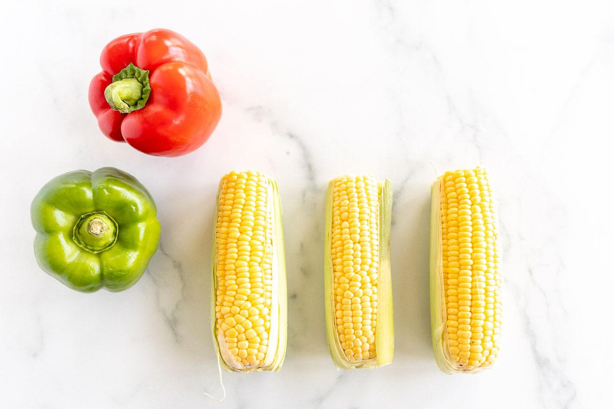 Corn on the cob and a red and green pepper, laid out on a white marble countertop
