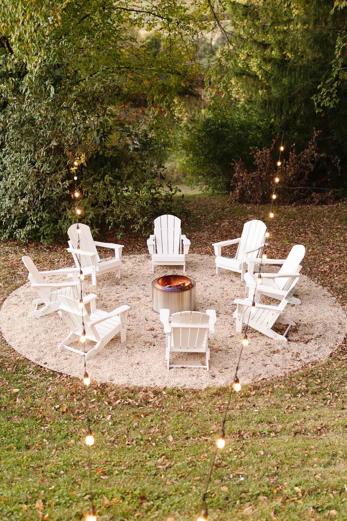 White Adirondack chairs, circling a Solo Stove on a DIY pea gravel fire pit patio. String lights are hung above, twinkling at dusk.