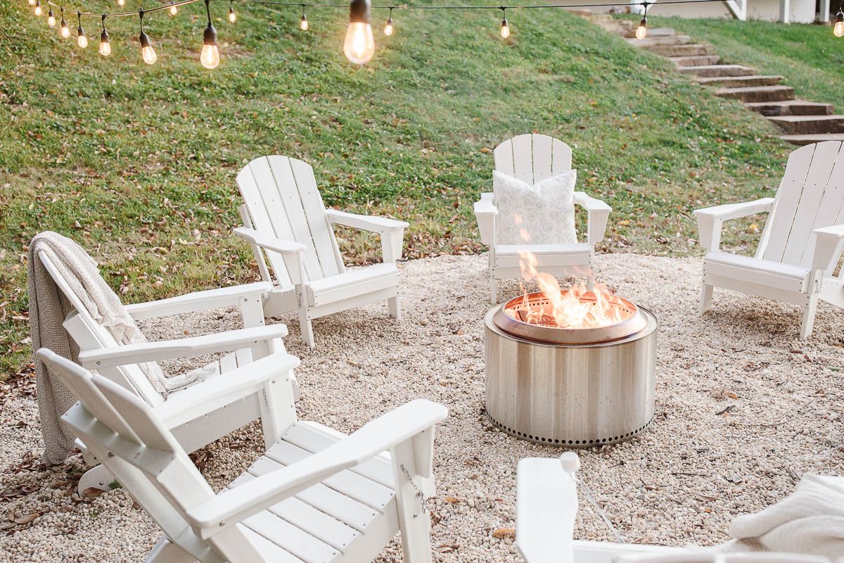 White Adirondack chairs, circling a Solo Stove on a DIY pea gravel fire pit patio. String lights are hung above, twinkling at dusk.