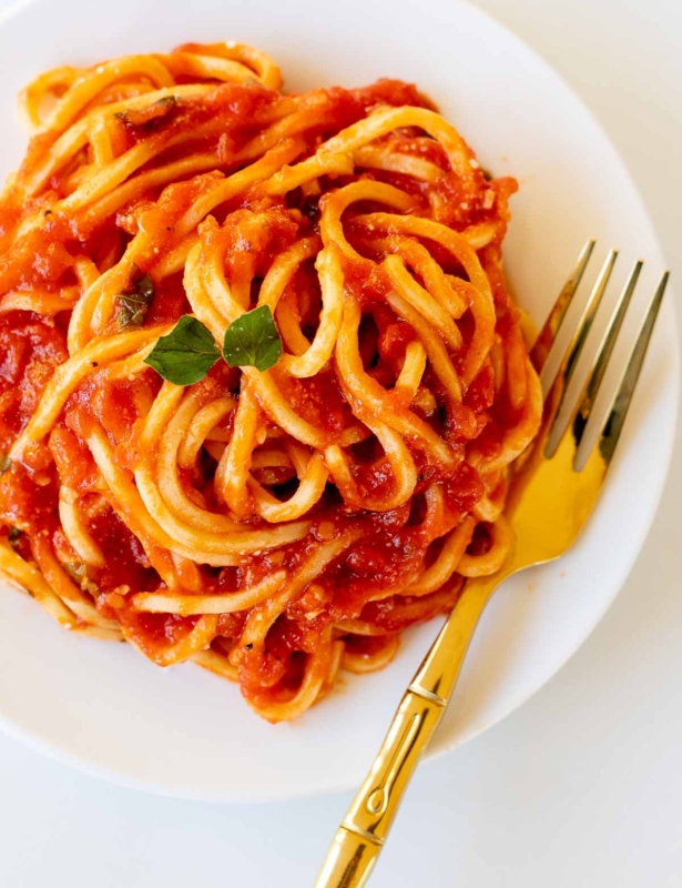 Spaghetti on a white plate, tossed in San Marzano tomato sauce, with a gold fork to the side.