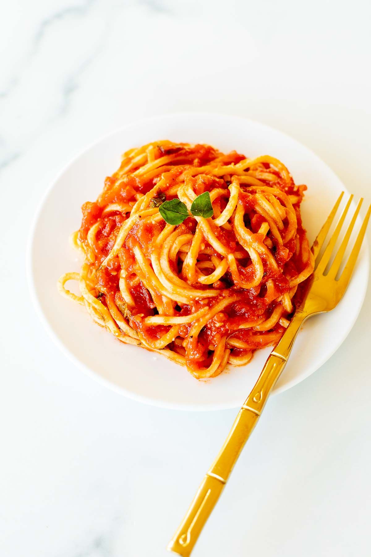 Spaghetti on a white plate, tossed in San Marzano tomato sauce, with a gold fork to the side.