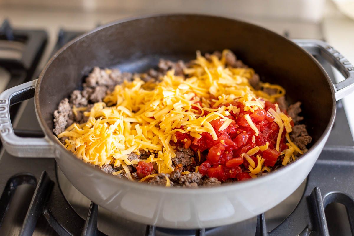 Taco meat in a gray pan on a stovetop, topped with tomatoes and cheese